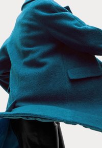 Abrigo de lana color verde azulado con cuello alto, mangas largas y un corte holgado; cuenta con un bolsillo frontal único y un acabado liso.