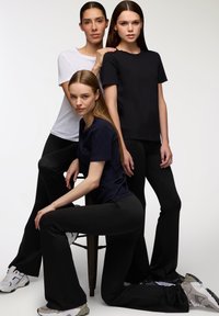 Tre donne vestite con pantaloni neri e t-shirt di colori solidi bianco, nero e blu scuro, posano davanti a uno sfondo leggero e semplice.