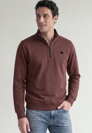 Homme aux cheveux foncés et bouclés, portant un sweat-shirt marron à fermeture éclair quart, et un jean bleu, debout avec une main dans la poche, regardant vers la gauche.