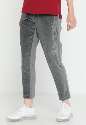 Person iført grå Champion joggingbukser, hvide Nike sneakers og en rød skjorte, stående mod en ensartet lys baggrund.