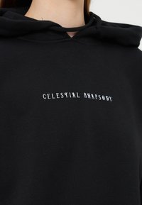 Zwarte hoodie met witte tekst "CELESTIAL RHAPSODY" gecentreerd op de borst, gedragen door een persoon die te zien is van nek tot bovenlichaam.