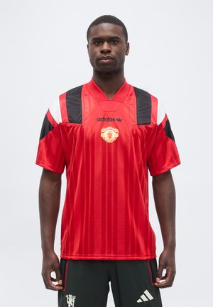 Man som bär en röd Manchester United Adidas fotbollströja med svarta och vita axelaccenter, ihop med svarta Adidas-shorts.