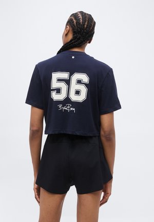 Person with braided hair wearing a navy cropped T-shirt with number 56 and signature on the back, paired with black shorts.