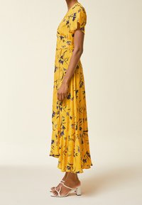 Żółta sukienka w kwiaty z krótkimi rękawami, dopasowanym gorsetem i rozkloszowanym dołem. Posiada niebieskie wzory kwiatowe oraz białe sandały na słupku.