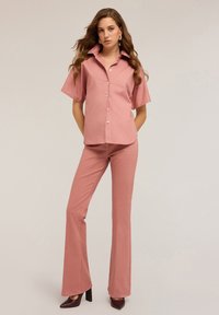 Una donna con lunghi capelli mossi indossa una camicia a maniche corte color mauve con bottoni, pantaloni svasati abbinati, orecchini dorati e tacchi alti marrone scuro.