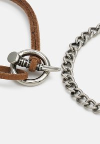 Brăţară din suede maro cu un închizător circular metalic şi o brăţară din lanţ de argint, cu zale strălucitoare şi o textură netedă.
