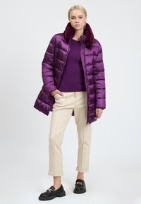 Frau steht mit einer lila Steppjacke mit Pelzkragen, lila Pullover, cremefarbenen Hosen und schwarzen Slippern mit goldenen Ketten.