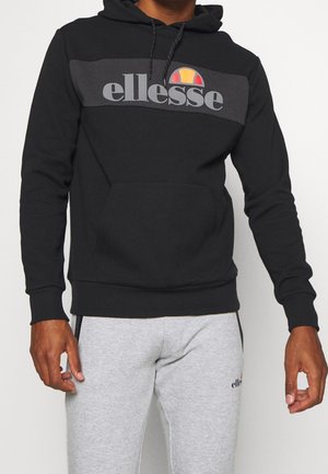 Mężczyzna w czarnej bluzie z kapturem ellesse z logo i jasnoszarych spodniach dresowych z małym logo ellesse na udzie.