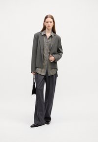 Grauer übergroßer Blazer über einem karierten Hemd mit Knöpfen, kombiniert mit lockeren dunklen Hosen und spitzen schwarzen Schuhen. Hält eine kleine schwarze Handtasche.