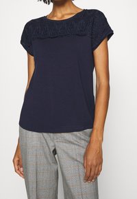 Žena v námořnickém modrém krátkém rukávu s krajkovým detailem na hrudi, doplněná šedými kostkovanými kalhotami.