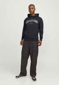 Sweatshirt à capuche noir avec le logo "JACK & JONES", poche avant, associé à un pantalon cargo noir ample et des chaussures de sport, sur un fond clair.