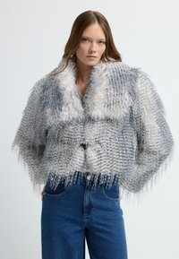 Grau-weiße füllige Kurzjacke aus Kunstfell mit strukturiertem Design, breitem Kragen und fransigen Abschlüssen, kombiniert mit blauen Jeans.