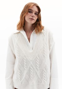 Gilet tricoté de couleur crème avec des motifs en relief, col en V et tissu texturé porté par-dessus une chemise blanche à col. Léger et ample.