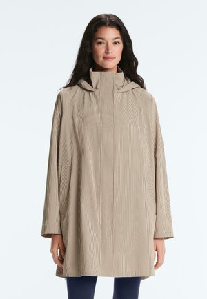 Nő bézs, nagy méretű, kutyanyelv mintás kabátban, magas gallérral és hosszú ujjakkal, sima háttér előtt áll.