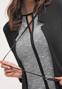 Černý kardigan s otevřeným předním dílem přes šedý top s kostkovaným vzorem. Má výstřih ve tvaru klíčové dírky a texturovaný design látky.