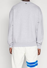 Personne portant un sweat-shirt gris clair avec un petit logo de crocodile vert au niveau de la nuque, assorti à un pantalon blanc avec deux bandes bleues.