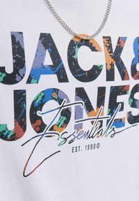 Hvid t-shirt med farverig tekst "JACK & JONES Essentials" i varierende fede skrifttyper, med en sølvkæde hængende over designet.