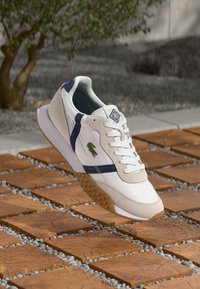 Witte sneakers met marineblauwe accenten, vetersluiting, gestructureerde beige neus, rubberen zool met patroon grip, en een klein groen logo aan de zijkant.