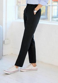 Černé tailored kalhoty s úzkým střihem, které mají boční kapsy, spárované s bílými plátěnými teniskami s červenými a modrými akcenty.