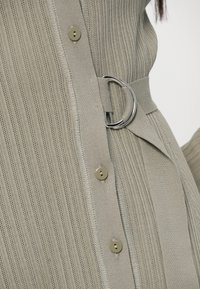 Lysegrå striktrøje med lodrette ribber, metalliske knapper og et stofbælte med en rund spænde til at stramme ind.