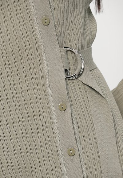 Haut gris clair en maille avec côtes verticales, boutons métalliques et ceinture en tissu avec boucle circulaire pour cintrer.