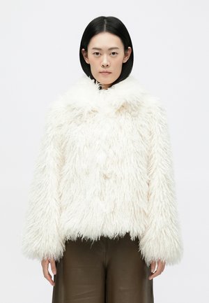 Bela jakna iz umetnega krzna z visokim ovratnikom, debelo teksturo in širokimi rokavi. Rob je skrajšan. Nosi se čez rjave hlače.