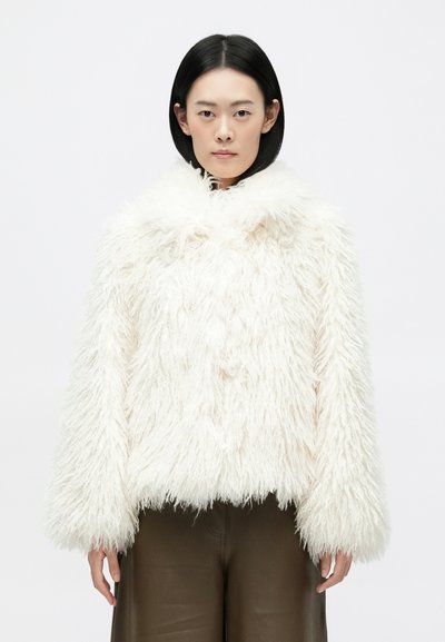 Chaqueta de pelo sintético blanca con un cuello alto, textura gruesa y mangas anchas. El dobladillo es corto. Usada sobre pantalones marrones.
