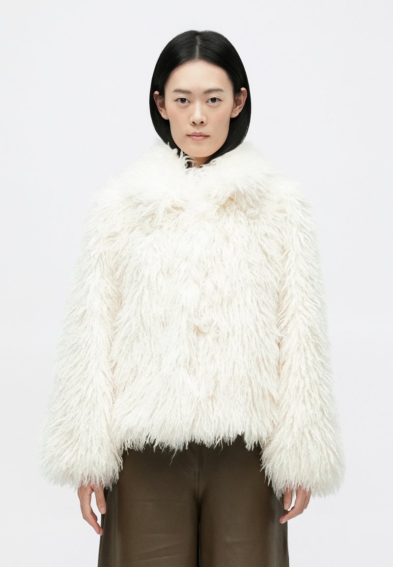 Biela kožušinová bunda s vysokým golierom, hustou textúrou a širokými rukávmi. Spodný okraj je skrátený. Nosená cez hnedé nohavice.
