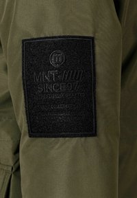 Olivgrön tygpatch på en jacka med präglad svart branding. Texten lyder "MNT SINCE 97 AUTHENTICALLY CRAFTED MNT COLLECTION."