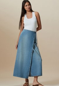 Jupe portefeuille en denim bleu clair à coupe droite, nouée à la taille, associée à un haut sans manches blanc orné de clous décoratifs.