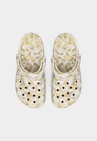 Ζευγάρι μπεζ και λευκών κλειστών παπουτσιών με σχέδιο παραλλαγής, με οπές αερισμού και ρυθμιζόμενα λουράκια στη φτέρνα, φωτογραφημένα από πάνω.