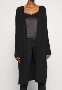 Long black cardigan made of soft knit material. Open front design with ribbed cuffs and side pockets. Worn over a satin black top.