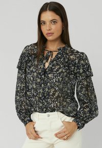 Femme aux longs cheveux bruns portant un blouson noir floral transparent avec des volants et un pantalon taille haute blanc, les mains dans les poches, sur fond uni.