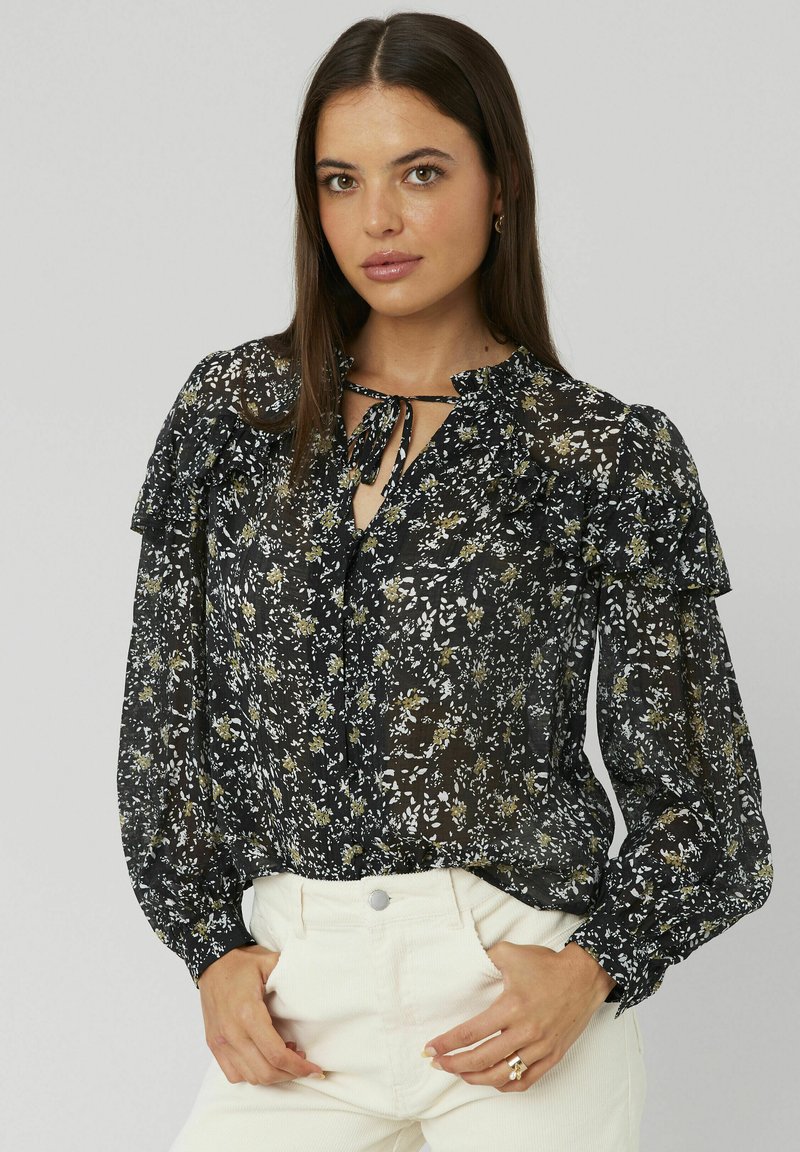 Femme aux longs cheveux bruns portant un blouson noir floral transparent avec des volants et un pantalon taille haute blanc, les mains dans les poches, sur fond uni.