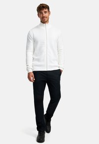 Weißer Zip-Jacke mit Zickzackmuster und gerippten Bündchen, kombiniert mit schwarzen Hosen und schwarzen Schuhen. Modell steht aufrecht vor einem neutralen Hintergrund.