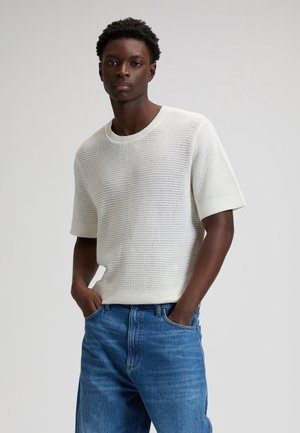 Hombre con suéter blanco de punto de manga corta y jeans azules, de pie con las manos en los bolsillos contra un fondo liso.