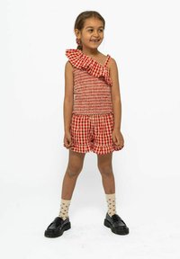 Monos con estampado de cuadros en rojo y blanco con un diseño de un hombro con volantes. Presenta pantalones cortos y zapatos negros con calcetines estampados.