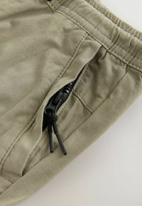 Pantaloni corti in tessuto morbido color beige chiaro, con una cintura elastica, dotati di una tasca laterale con zip e un cordino per la regolazione.