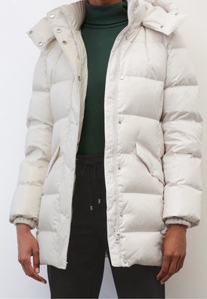 Person iført en beige quiltet vinterjakke over en grøn rullekravetrøje og sorte bukser, stående foran en ensfarvet baggrund.