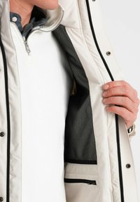 Giacca esterna beige con una cerniera argento, che rivela una fodera in rete nera e uno strato interno bianco. Presenta una tasca con cerniera all'interno.