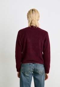 Burgundi värvi fliisist sviiter, millel on tekstuuriga pind, soonikkoes kaelus ja pikad varrukad, kombineeritud kõrge vöökohaga siniste teksadega.