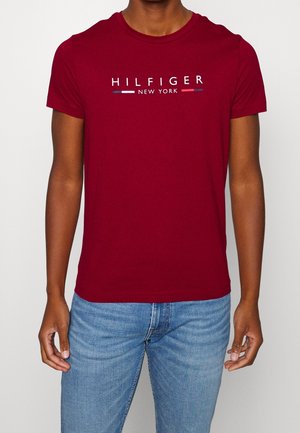 Homme portant un t-shirt rouge à manches courtes avec le texte "HILFIGER NEW YORK" et des bandes de couleurs, assorti à un jean bleu clair.