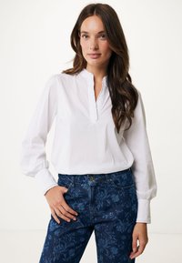 Blusa bianca a maniche lunghe con colletto volant e polsini elastici, abbinata a jeans in denim scuro con motivo floreale.