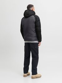 Schwarze und graue Steppjacke mit Kapuze, kombiniert mit dunklen Cargo-Hosen und beigefarbenem Schuhwerk. Glatte Oberfläche mit matter Verarbeitung.