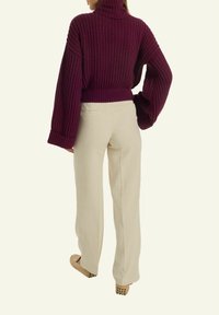 Person, die einen voluminösen lila Rollkragenpullover, beige gerade geschnittene Hosen und beige Slipper mit Gummigriffen trägt, steht mit dem Rücken zur Kamera.