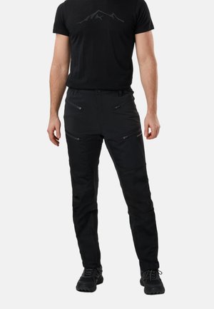 Mand iført sorte vandrebukser med lynlåslommer og sorte sko, stående foran en ensfarvet lys baggrund.