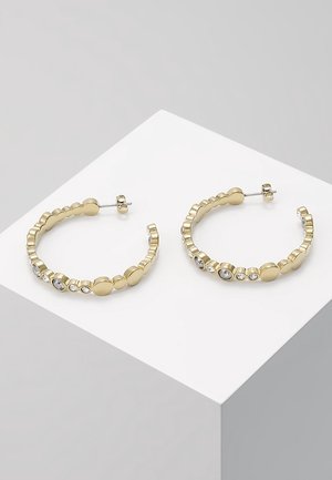 Boucles d'oreilles créoles dorées ornées de pierres précieuses claires rondes et ovales, présentées sur un cube blanc sur un fond gris clair.