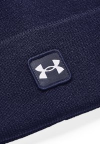 Bonnet en tricot bleu marine avec un patch carré portant le logo Under Armour en blanc. Texture douce avec un ajustement serré et des détails en côtes fines.