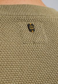 Olivgrüner Strickpullover mit texturiertem Oberflächen, ausgestattet mit einem schwarzen Etikett mit gelber Schrift und einem runden Akzent am Hals.