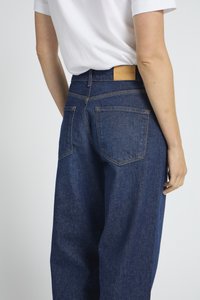 Osoba odjevena u tamnoplave široke traperice i bijelu košulju s kratkim rukavima uvučenu unutra, prikazana od struka unazad do sredine bedara.